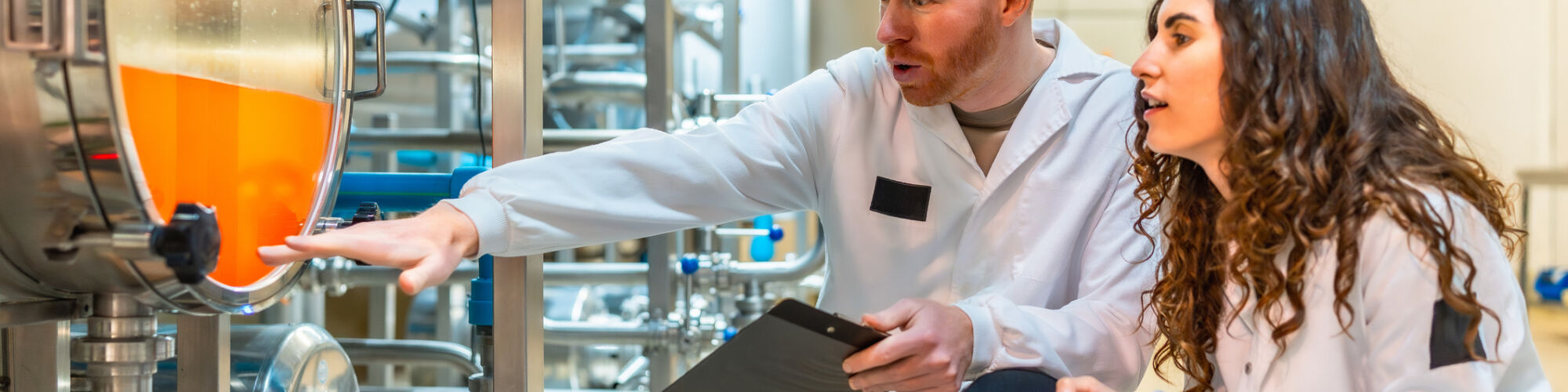Two engineers wearing lab coats inspecting the quality and production levels of beer inside a large stainless steel fermenting tank at a modern beverage factory